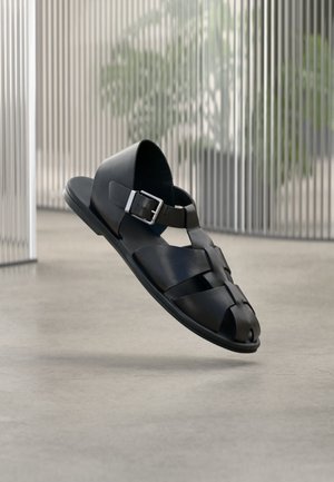 Schwarze Leder-Herrensandale mit silberner Schnalle, offenem Zehendesign, schwebend über einem glatten Betonboden.