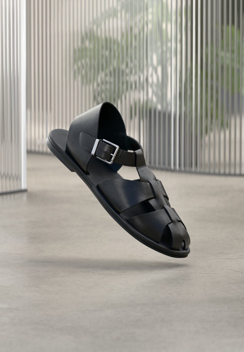 Μαύρο ανδρικό δερμάτινο σανδάλι με ασημένια αγκράφα, σχέδιο ανοιχτής μύτης, πλέει πάνω από ομαλό τσιμεντένιο πάτωμα.