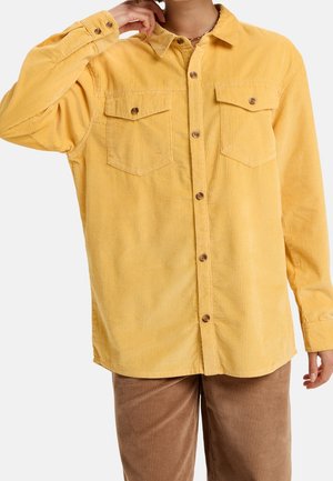 Homme portant une chemise jaune en velours côtelé à manches longues avec des poches poitrine à boutons et un pantalon en velours côtelé marron, la main touchant le cou.