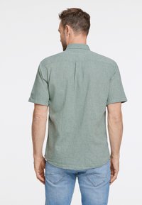 Camicia a maniche corte con colletto in tessuto verde chiaro con finitura texture. Presenta uno yoke posteriore e un orlo leggermente curvato. Indossata con jeans blu.