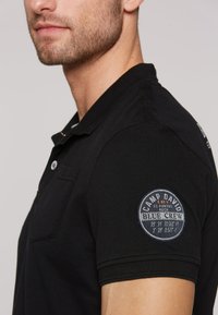 Maglietta polo nera con finitura testurizzata, dotata di colletto, due bottoni e una toppa circolare sulla manica sinistra con cuciture bianche e testo.