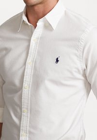 Witte overhemden gemaakt van katoen, met een kraag, knopen aan de voorkant en een klein marineblauw geborduurd logo op de linkerborst.