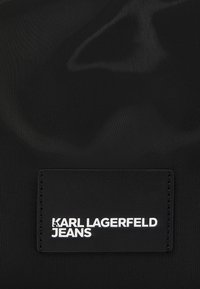 Μαύρο νάιλον ύφασμα με υφή. Περιλαμβάνει μια ορθογώνια μαύρη ετικέτα με την ένδειξη "KARL LAGERFELD JEANS" σε λευκά γράμματα.