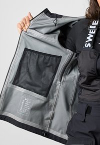 Vedenpitävän takin sisäpuoli harmaasta kankaasta, jossa musta verkkotasku. Ominaisuuksiin kuuluu hoito-ohjetarra ja ripustuslenkki. Vetoketjullinen suljenta.