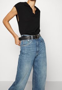 Schwarzes ärmelloses Poloshirt, kombiniert mit blauen weit geschnittenen Jeans. Das Top hat einen Kragen und eine lockere Passform; die Jeans besitzen ein klassisches Fünf-Taschen-Design.