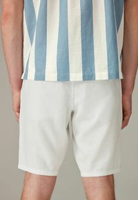 Cămașă cu mânecă scurtă, cu dungi albastre și albe, având un model texturat; asortată cu pantaloni scurți albi, până la genunchi, dintr-un material ușor.