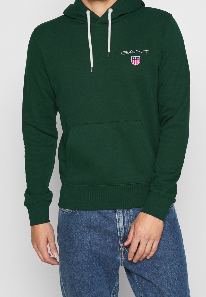 Sweat à capuche en coton vert avec une poche avant, une capuche à cordon de serrage et le logo GANT. Comprend des poignets et un ourlet côtelés. Associé à un jean en denim bleu.
