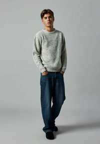 Helles graues geripptes Strickpullover mit Rundhalsausschnitt, kombiniert mit lockeren dunkelblauen Jeans. Das Model steht mit den Händen in den Taschen.