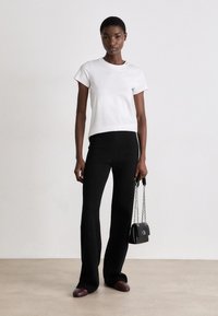T-shirt bianca aderente, pantaloni neri a costine svasati, flats bordeaux e una piccola borsa nera con tracolla a catena, il tutto su uno sfondo neutro.