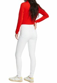 Jeans skinny blancos con bolsillos traseros, combinados con una blusa ajustada de manga larga roja. Zapatillas blancas con detalles amarillos completan el atuendo.