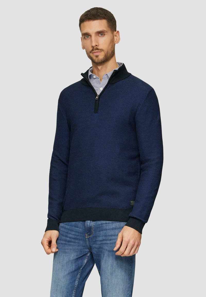 Marinefarbener Pullover mit Reißverschluss, strukturiertem Strick, kontrastierendem Kragen sowie gerippten Bündchen und Saum; über einem gestreiften Hemd getragen; kombiniert mit hellblauen Jeans.