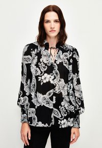 Schwarze Bluse mit weißen Paisley- und Blumenmuster, ausgestattet mit einem hohen Kragen mit Krawatte, gerüschten Schultern und langen Puffärmel.