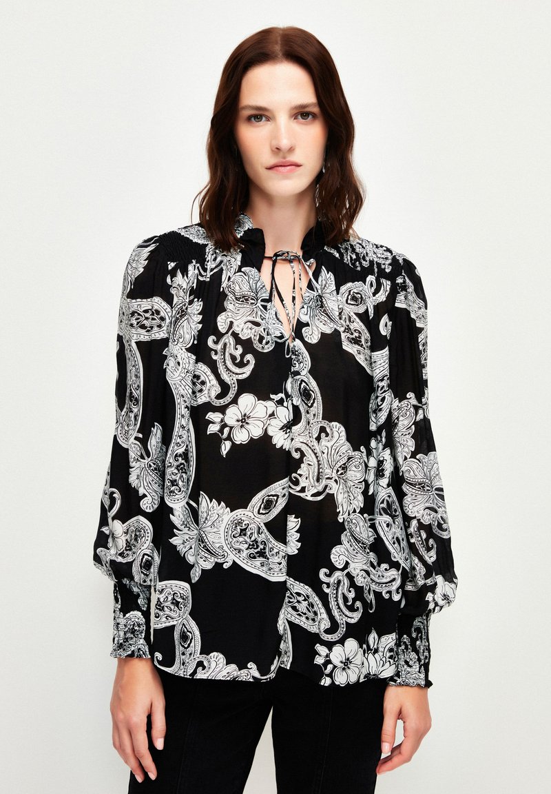 Schwarze Bluse mit weißen Paisley- und Blumenmuster, ausgestattet mit einem hohen Kragen mit Krawatte, gerüschten Schultern und langen Puffärmel.