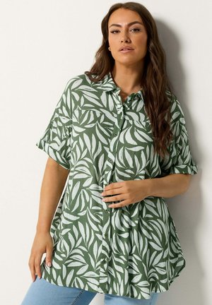 Frau mit langen braunen Haaren trägt ein lockeres grünes Hemd mit weißen Blattmustern und hellblaue Jeans, posiert vor einer weißen Wand.