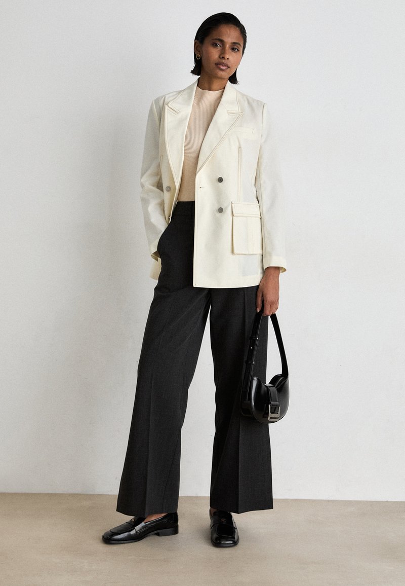 Doppelt gefütterte cremefarbene Blazer mit Schulterpolstern, kombiniert mit grauen, weitgeschnittenen Hosen. Schwarze Loafers und eine strukturierte schwarze Tasche runden den Look ab.