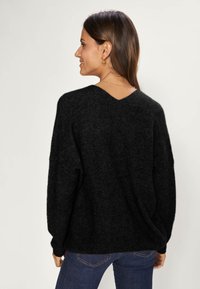 Zwarte gebreide trui met een V-hals en verlaagde schouders. Gestructureerde stof en een casual pasvorm. Draagt met donkerblauwe jeans, gezien vanuit de rug.