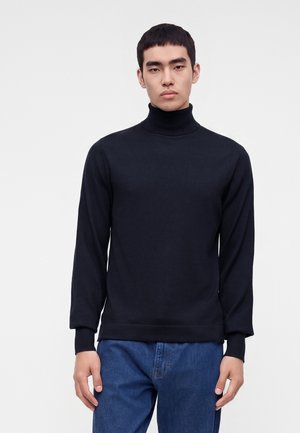 Jeune homme aux cheveux courts et noirs portant un pull noir à col roulé et un jean bleu, debout devant un fond blanc uni.