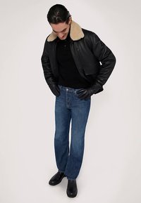 Svart läderbomberjacka med beige pälskrage, svart pikétröja, blå jeans och svarta stövlar; handskar är på, händerna vilar på låren.