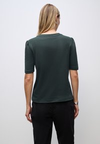Žena s dlouhými blond vlasy, oblečená v tmavě zeleném tričku s krátkým rukávem a černých kalhotách, viděná zezadu na jednolitém světlém pozadí.