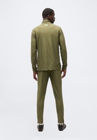 Homme debout de dos, portant un survêtement vert olive à manches longues, pantalon, et des chaussures de sport noires sur un fond blanc uni.