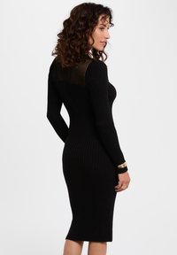 Robe noire à rayures avec manches longues, design ajusté et panneau arrière transparent à rayures ; longueur genou avec tissu texturé. Bracelets en or au poignet.