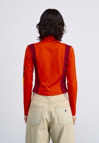 Langermet topp i klar oransje stoff med burgunder sidesømmer og høy krage; kombinert med lyse beige, løstsittende bukser.