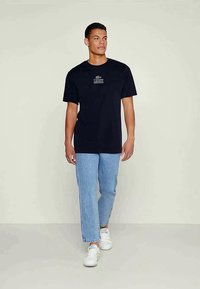 Camiseta de algodón azul marino de manga corta, con un pequeño logo en el pecho. Lleva puesta con jeans ajustados de color azul claro y zapatillas blancas.