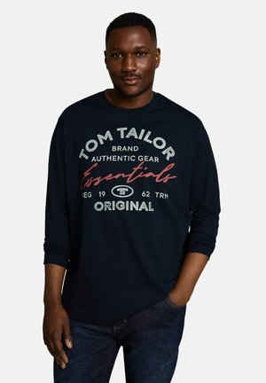 Junger Mann, der ein dunkelblaues Langarmshirt mit dem Text "Tom Tailor Authentic Gear Essentials Original" trägt, steht vor einem schlichten hellen Hintergrund.