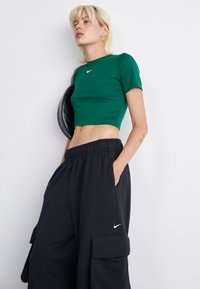 Henkilö, jolla on päällään vihreä lyhyt Nike-paita, mustat Nike-haremihousut ja joka kantaa mustaa olkalaukkua, seisoo valkoista taustaa vasten.