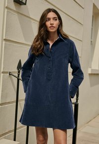 Granatowa sukienka koszulowa z bawełny, z długimi rękawami, z zapinaniem na guziki i kieszeniami naszytymi. Miękka w dotyku, z klasycznym kołnierzykiem i luźnym krojem.