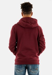 Bordeauxrode hoodie met een trekkoordkap, lange mouwen en een kangoeroezak. De achterkant is glad, gecombineerd met blauwe jeans.