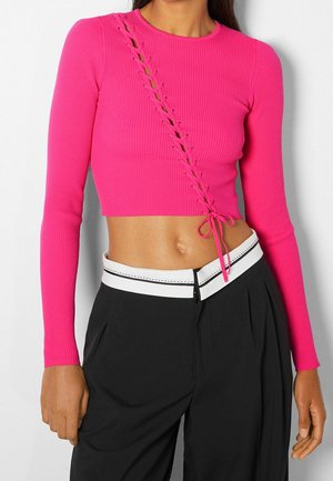 Femme portant un crop top à manches longues rose vif avec un détail lacé en diagonale et un pantalon noir avec une ceinture blanche à la taille.