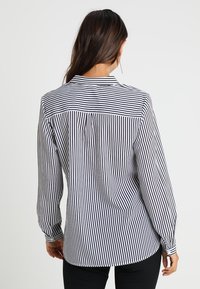 Blouse rayée noir et blanc avec un col rabattable, des manches longues et un détail plis à l'arrière. Le tissu semble lisse et léger.