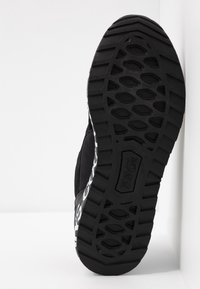 Svart skosula med texturerade mönster och "Versace Jeans Couture"-logotypen präglad i centrum, vinklad mot en vit bakgrund.