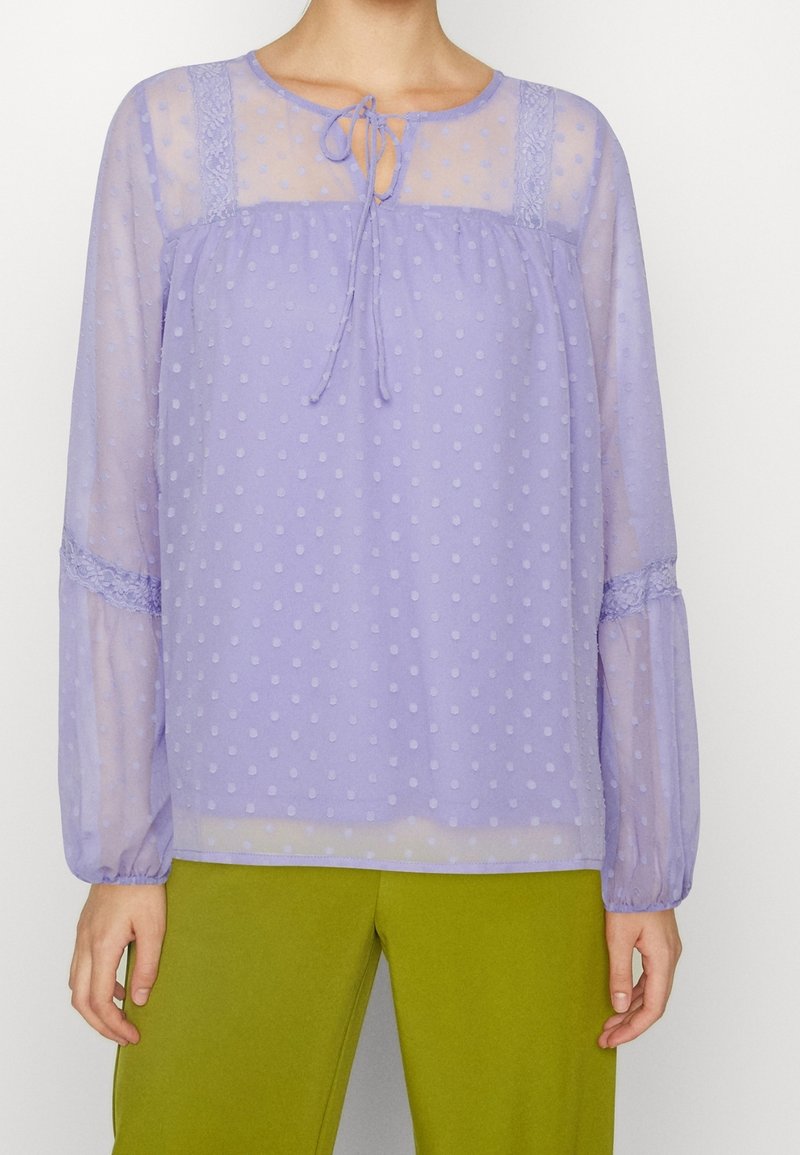 Blouse lilas clair en tissu transparent, avec un motif à pois et un détail en lien au niveau du col. Elle présente de longues manches froncées avec des accents en dentelle.