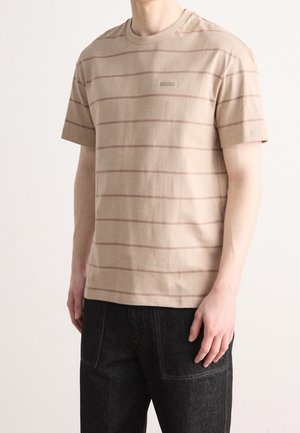 Camiseta de algodón beige con rayas horizontales en marrón claro, mangas cortas y cuello redondo. Parche de logo notable en el pecho.