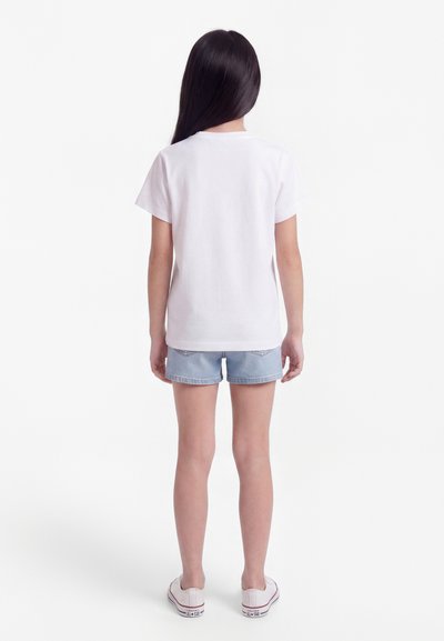 Κορίτσι με μακριά σκούρα μαλλιά, ντυμένο με λευκό μπλουζάκι, ανοιχτό μπλε τζιν σορτς και λευκά αθλητικά παπούτσια, στέκεται με την πλάτη γυρισμένη σε λευκό φόντο.