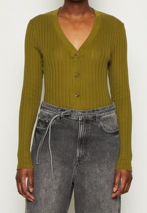 Cardigan a coste verde oliva con scollatura a V, caratterizzato da tre bottoni marroni sul davanti. Abbinato a jeans neri scoloriti a vita alta.