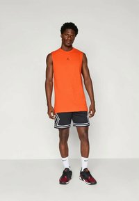 Oranž särk ilma varrukateta spordiriietuses, millel on must logo, koos mustade lühikeste pükstega, millel on valge äär. Jalatsiteks on mustad ja punased sportlikud tossud.