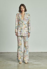 Vrouw staat in een op bloemenpatroon gesneden blazer en wijde broek, beige puntige schoenen, neutrale achtergrond.