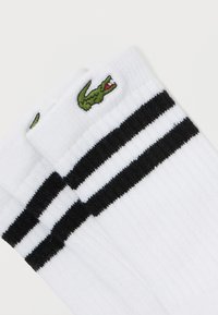Calcetines de pibón blancos con textura acanalada, que cuentan con dos rayas negras y un logo de cocodrilo verde en el puño. Material suave de algodón.