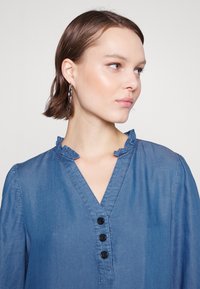 Blouse en denim bleu clair avec un décolleté en V orné de volants, manches longues et un patte de boutonnage à quatre boutons noirs. Texture lisse.