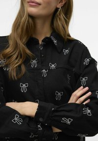 Zwarte shirt met witte geborduurde strikjes, knoopsluiting, kraag en geplooide manchetten. Gladde textuur met zichtbare stikdetails.