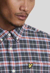 Camicia a quadri in navy, rosso e azzurro, realizzata in un tessuto morbido. Presenta bottoni bianchi e un piccolo logo ricamato giallo.