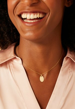 Collier en chaîne dorée avec un petit pendentif ovale. La chaîne est délicate et le pendentif a une surface lisse et réfléchissante. Porté sur un haut de couleur rose pâle.