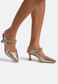 Mules metálicos plateados sin cierre, con punteras puntiagudas, tacones de bloque bajos y correas adornadas con piedras transparentes.