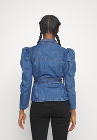 Džínová bunda s vypouklými rameny, s páskem v pase a detailem v oblasti zad. Má středně modrou barvu a klasický límeček. Viditelné černé kalhoty.