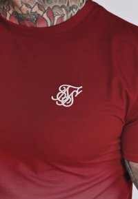 Camiseta roja de manga corta con un logo bordado en blanco en el lado izquierdo del pecho. Tejido suave con una textura ligeramente degradada.