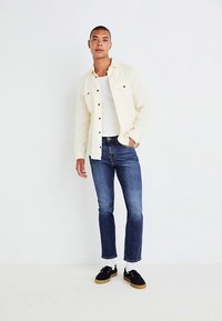 Camisa de botão creme claro sobre uma t-shirt branca, combinada com jeans azul escuro e sapatos pretos com sola de borracha. Design simples, ajuste casual.