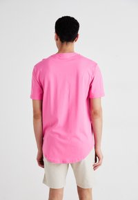 Trumpomis rankovėmis ryškiai rožinės medvilnės marškinėliai su apvalia iškirpte ir ilgesniu galine apačia, derinami su šviesiai smėlio spalvos šortais.